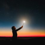 A silhouette of a person holding a glowing wand under a vibrant sunset sky, creating a magical atmosphere.